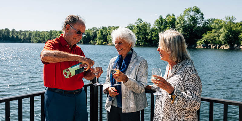 one older man and two women with gray hair talking and holding wine glasses on a fenced patio with woods and a lake in the background