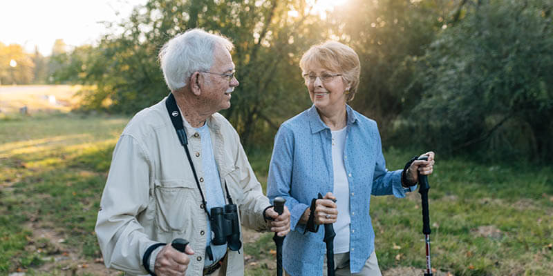 an older man and woman on a hike, both with walking sticks and man with binoculars around his neck