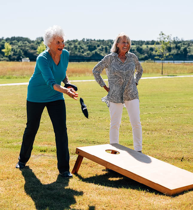 two older women outside in recreational area smiling and playing baggo game together